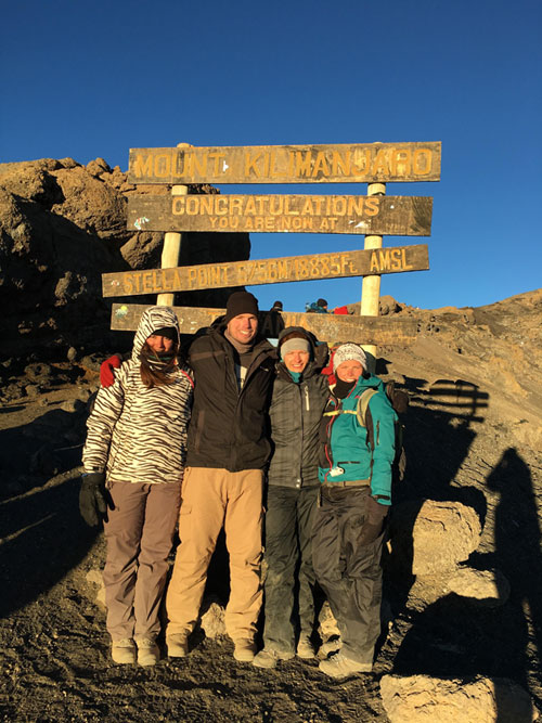Four hikers pose in front of Mount Kilimanjaro sign